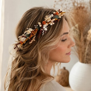 Op de afbeelding: Een bloemenkrans met gedroogde bloemen in tinten oranje, crème en groen. De krans wordt op het hoofd gedragen en heeft een delicate, natuurlijke uitstraling. Het haaraccessoire bevat een kanten lint.