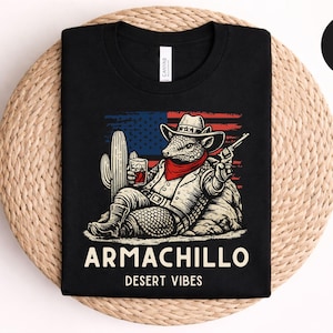 May include: Black t-shirt with a graphic of an armadillo in a cowboy hat, red bandana, and a gun, with a cactus and American flag background. The text "Armachillo Desert Vibes" is printed below the graphic.