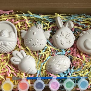 May include: An Easter craft kit featuring unpainted plaster figures: a basket of eggs, a chick, a bunny, a chick, a carrot, and an egg. The kit includes paint pots and a paintbrush, presented in a box with colourful paper shreds.
