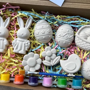 May include: An Easter-themed paint kit featuring white bunny and egg figurines, a chick, and a flower. The kit includes a paintbrush and a selection of colourful paints in small containers, all in a box with colourful paper filler.