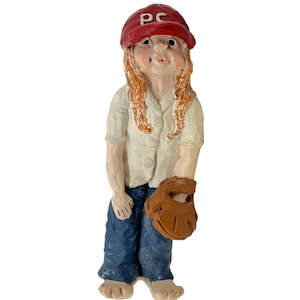 May include: A ceramic figurine of a person in a baseball uniform. The figure wears a red cap with "PC" on it, a white shirt, blue jeans, and a brown baseball glove. The figurine has red hair.
