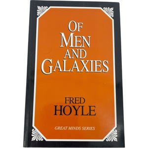 May include: A hardcover book titled Of Men and Galaxies by Fred Hoyle from the Great Minds Series. The cover features a bold orange rectangular center panel with white text, framed by a black border with decorative white corner flourishes.