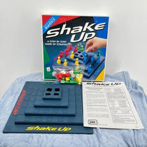 May include: A boxed board game titled "Shake Up" with a blue and green box. The game includes a blue game board, dice, and colorful game pieces. The box reads "A Step by Step Game of Strategy."