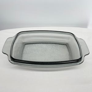 May include: A rectangular, clear glass baking dish with rounded corners and two handles. The dish has a slightly smoky gray tint and is designed for oven use. The overall shape is a classic, functional design for baking and serving.