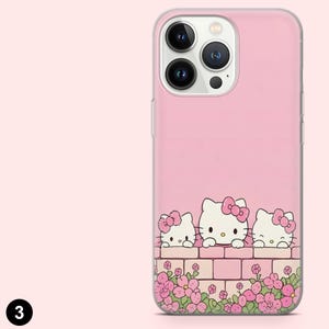 May include: Pink phone case featuring three Hello Kitty characters peeking over a brick wall with pink flowers. The case has a clear border and a pink background. The phone's camera lenses are visible.