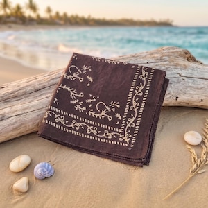 K&ouml;nnte beinhalten: Ein dunkelbraunes Bandana mit cremefarbenem Blumen- und Rankenmuster, gefaltet und auf einem St&uuml;ck Treibholz an einem Sandstrand liegend. Das Bandana ist quadratisch. Kleine Muscheln und eine Weizen&auml;hre liegen daneben.