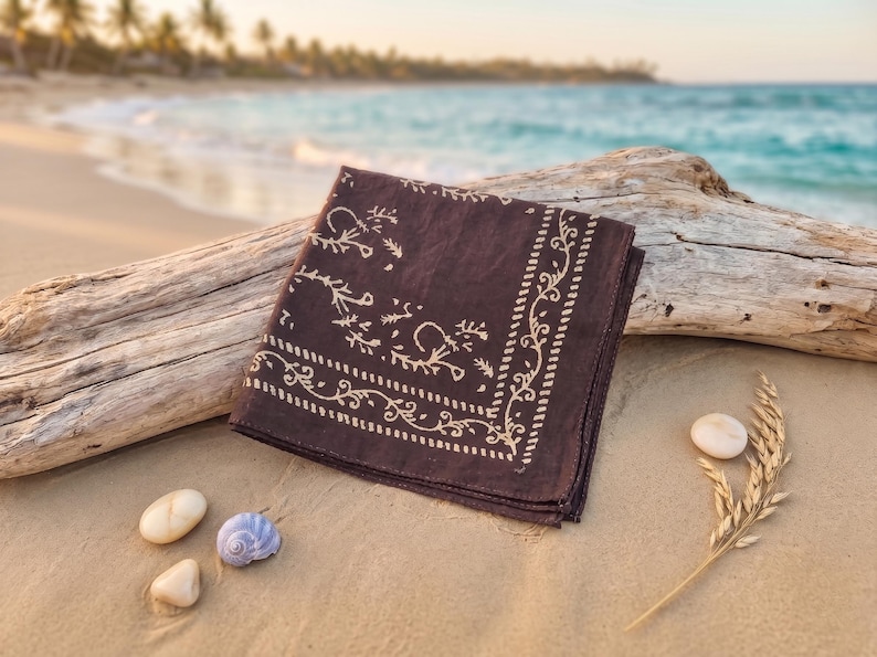 K&ouml;nnte beinhalten: Ein dunkelbraunes Bandana mit cremefarbenem Blumen- und Rankenmuster, gefaltet und auf einem St&uuml;ck Treibholz an einem Sandstrand liegend. Das Bandana ist quadratisch. Kleine Muscheln und eine Weizen&auml;hre liegen daneben.