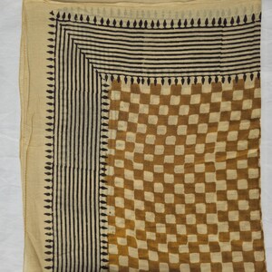 May include: A beige scarf with a geometric pattern. The scarf features a checkerboard design in shades of brown and beige, with a border of vertical black lines and a row of black teardrop shapes.