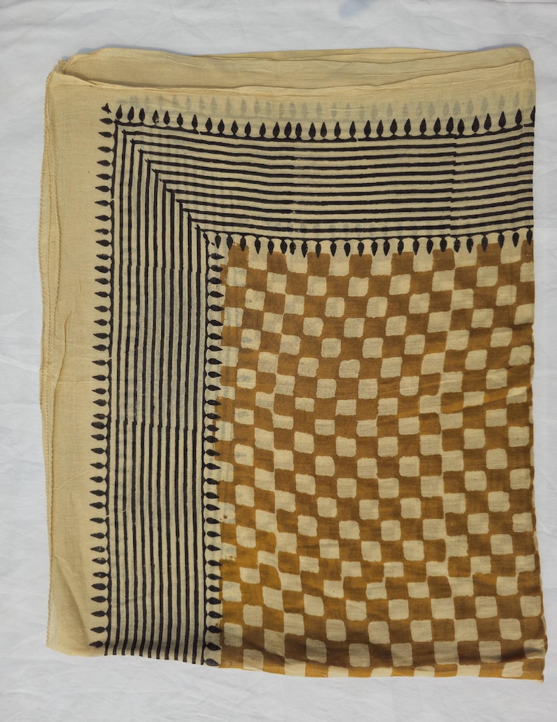 May include: A beige scarf with a geometric pattern. The scarf features a checkerboard design in shades of brown and beige, with a border of vertical black lines and a row of black teardrop shapes.
