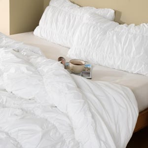 May include: A white bed set with a textured design. The set includes a duvet and two pillowcases. A cup of coffee and a magazine rest on the bed. The bed is made up with a neutral-colored fitted sheet.
