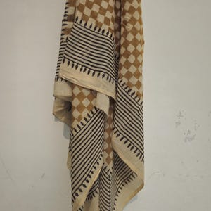 May include: A tan and cream scarf with a geometric pattern. The scarf features a repeating diamond pattern in tan and cream, with black and cream striped borders and decorative edges. The scarf is draped against a white wall.