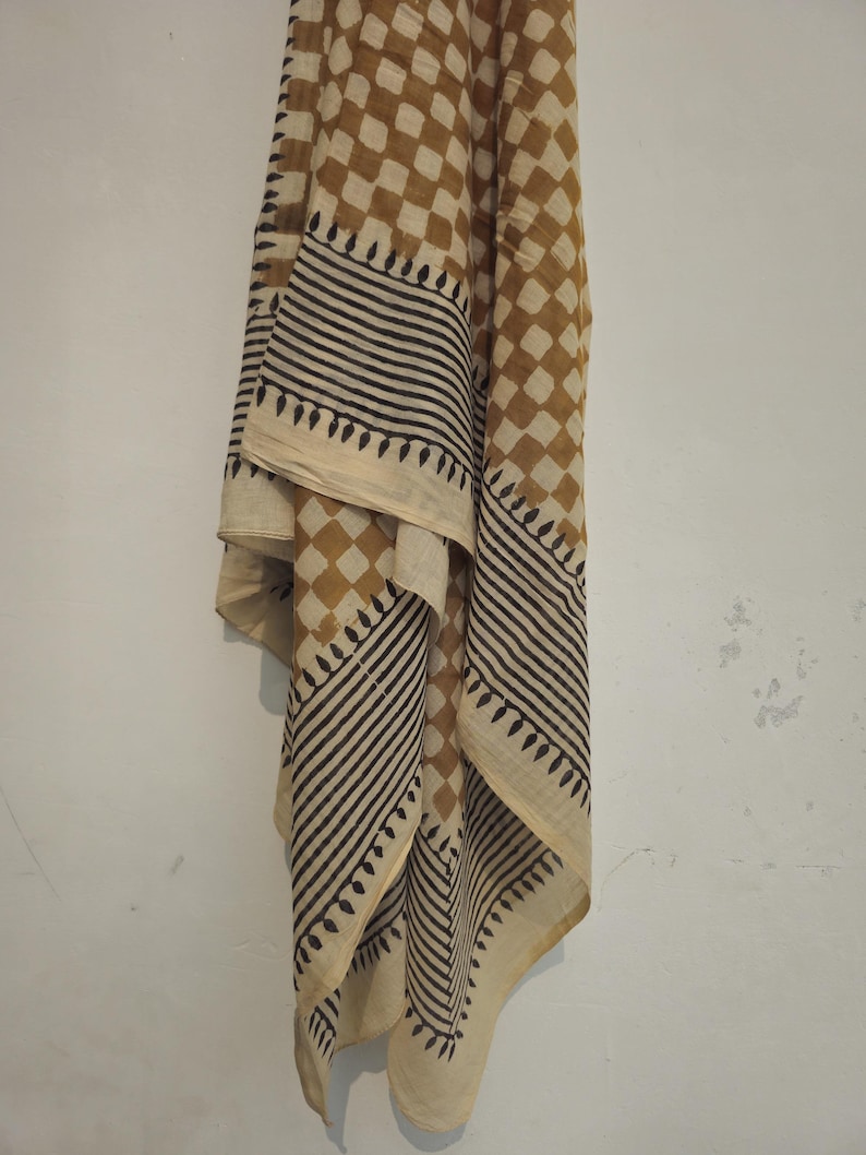 May include: A tan and cream scarf with a geometric pattern. The scarf features a repeating diamond pattern in tan and cream, with black and cream striped borders and decorative edges. The scarf is draped against a white wall.