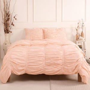 May include: A peach-colored bed set featuring a ruched design. The set includes a comforter and two pillow shams. The bedding is displayed on a wooden bed frame against a white wall. Decorative vases and a small table are visible.