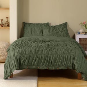 May include: An olive green bed set with a ruched design. The set includes a duvet cover and two pillowcases. The duvet cover has a buttoned edge. The bedding is displayed on a wooden bed frame.