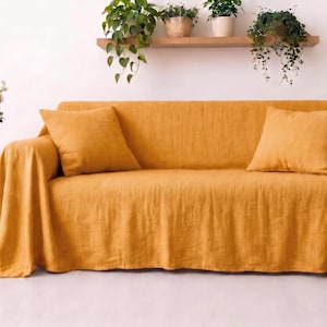 May include: A mustard yellow sofa cover draped over a couch, featuring two matching square throw pillows. The fabric has a visible textured weave, and the cover extends over the arms and down to the floor, creating a relaxed, layered appearance.