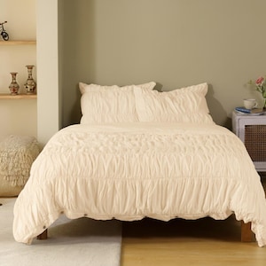 May include: A cream-colored bed with a ruched duvet cover and matching pillow shams. The bed is set against a neutral-toned wall, with a small side table and decorative items visible in the background. The bed frame is made of wood.