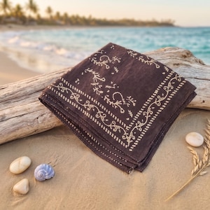 K&ouml;nnte beinhalten: Ein gefaltetes dunkelbraunes Bandana mit cremefarbenem Blumenmuster liegt an einem Sandstrand. Das Bandana liegt neben Treibholz, Muscheln und einem Weizen&auml;hrenzweig. Das Meer und Palmen sind im Hintergrund zu sehen.