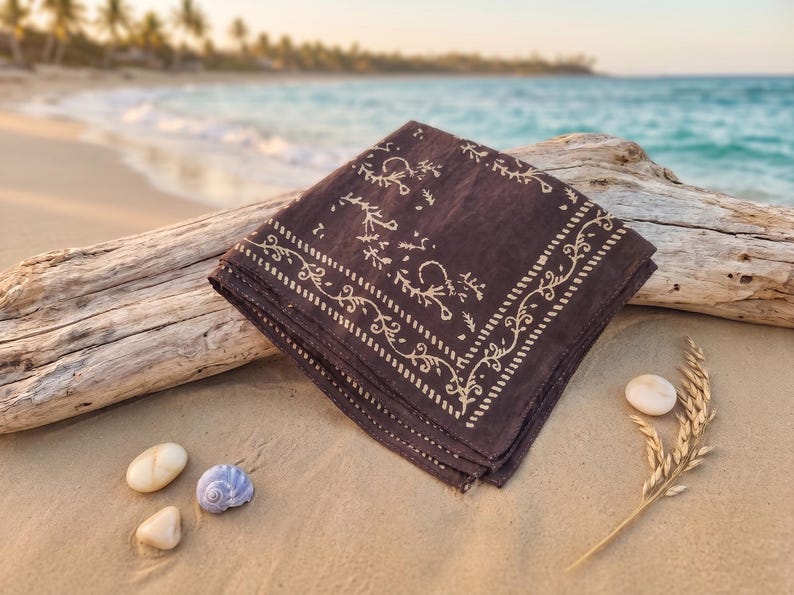 K&ouml;nnte beinhalten: Ein gefaltetes dunkelbraunes Bandana mit cremefarbenem Blumenmuster liegt an einem Sandstrand. Das Bandana liegt neben Treibholz, Muscheln und einem Weizen&auml;hrenzweig. Das Meer und Palmen sind im Hintergrund zu sehen.