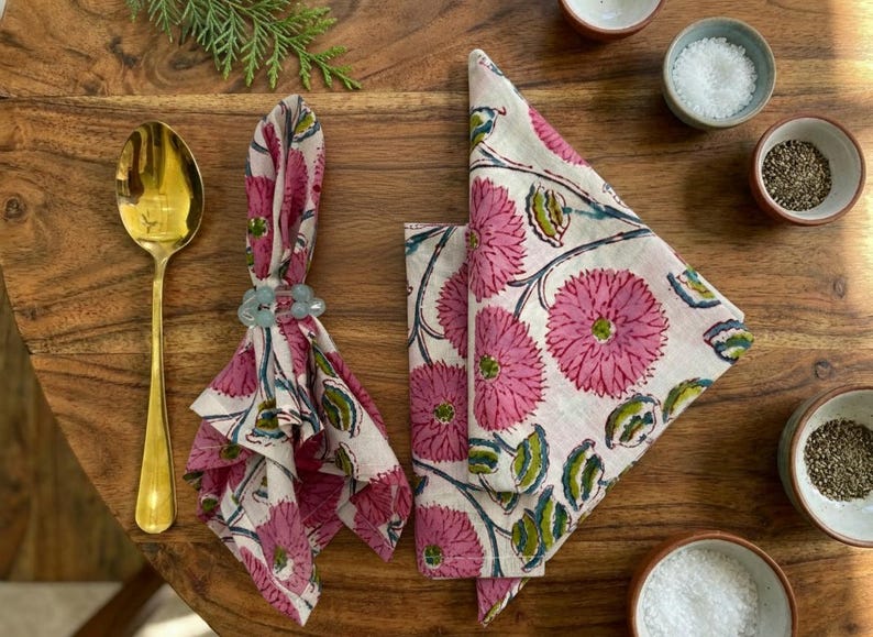 May include: A dining table arrangement with pink and white floral napkins, a gold spoon, and small bowls of salt and pepper. The napkins are folded and secured with a blue napkin ring. A sprig of greenery adds a natural touch.