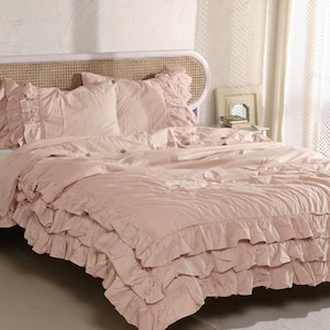 May include: A blush pink bed set with ruffled detailing. The set includes a comforter, pillow shams, and a bed skirt. The headboard is a light-colored wicker design. The bedding is made of a soft, textured fabric.