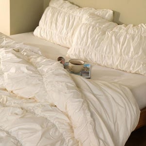 May include: A bed with white bedding, including a comforter and two ruffled pillows. A cup of coffee and a magazine rest on the bed. The room has a neutral color palette.