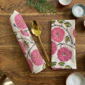 May include: Two floral print napkins, one rolled, and a gold spoon on a wooden table. The napkins display a pink flower design with green leaves and blue stems. Small bowls of salt and pepper are also visible.