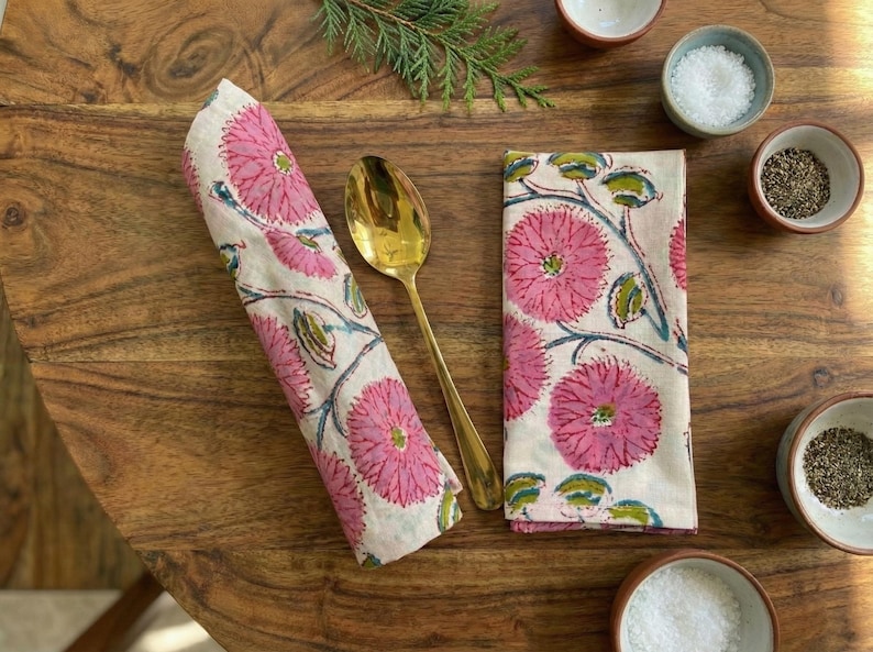 May include: Two floral print napkins, one rolled, and a gold spoon on a wooden table. The napkins display a pink flower design with green leaves and blue stems. Small bowls of salt and pepper are also visible.