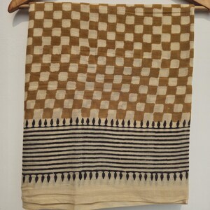 May include: A tan and brown checkered fabric scarf hanging on a wooden hanger. The scarf has a repeating pattern of brown squares on a tan background. The bottom of the scarf has a border of black horizontal lines and teardrop shapes.