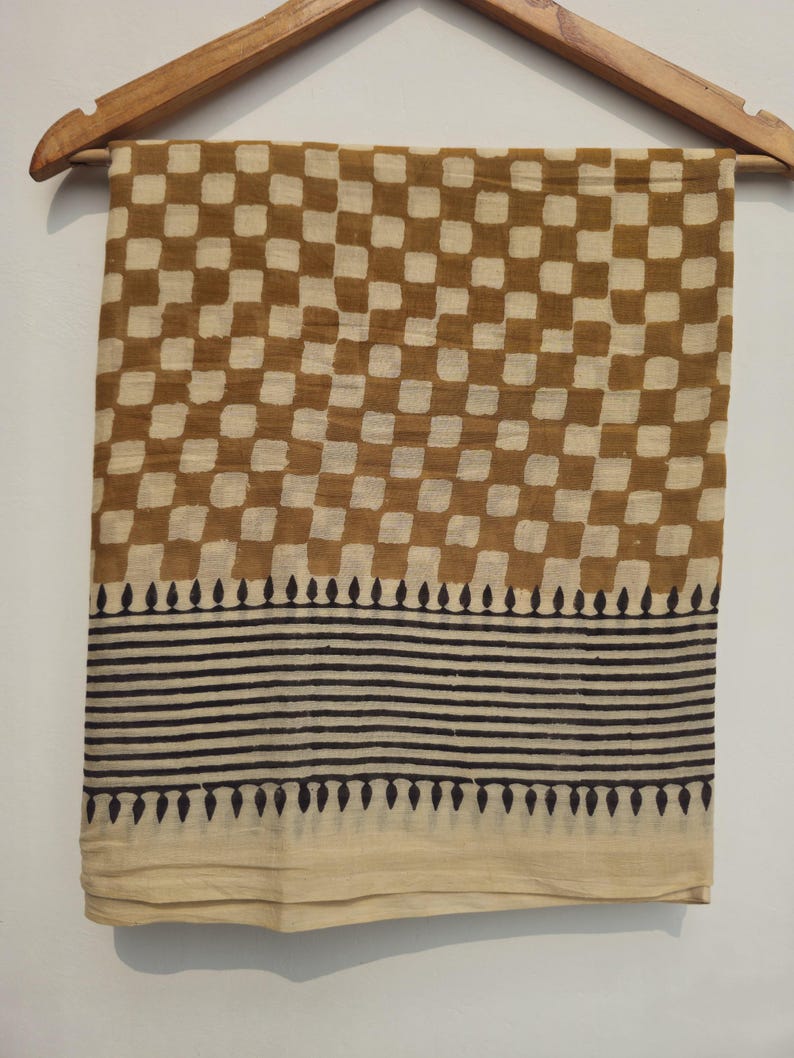 May include: A tan and brown checkered fabric scarf hanging on a wooden hanger. The scarf has a repeating pattern of brown squares on a tan background. The bottom of the scarf has a border of black horizontal lines and teardrop shapes.