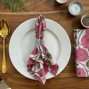 May include: A table setting featuring a white plate, gold cutlery, and a folded napkin with a pink floral design. A napkin ring with a blue flower design holds the napkin. Small bowls of salt and pepper are also present.