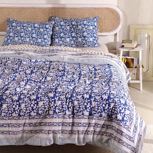May include: A blue and white floral patterned quilt set with two matching pillowcases. The bedding features a repeating floral design and a striped border. The headboard is made of woven rattan.