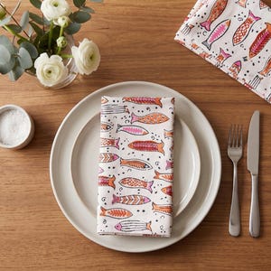 May include: A folded white napkin with a colorful fish pattern rests on a white plate, next to a fork and knife. A small bowl of salt and a vase of white flowers are also on the wooden table. The napkin has orange, pink, and black fish.