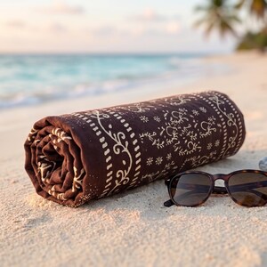 K&ouml;nnte beinhalten: Ein gerolltes braunes Strandtuch mit wei&szlig;em Blumenmuster und eine Sonnenbrille mit Schildpattmuster liegen auf einem Sandstrand. Das Meer und Palmen sind im Hintergrund zu sehen.