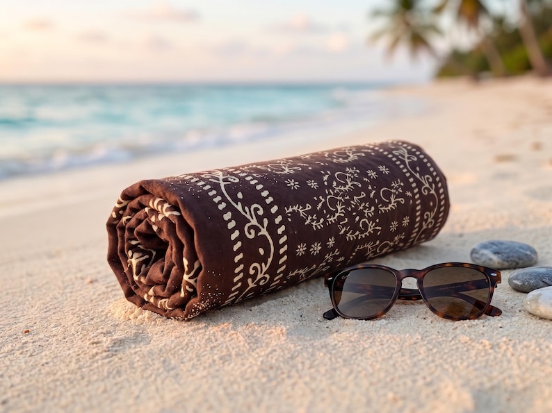K&ouml;nnte beinhalten: Ein gerolltes braunes Strandtuch mit wei&szlig;em Blumenmuster und eine Sonnenbrille mit Schildpattmuster liegen auf einem Sandstrand. Das Meer und Palmen sind im Hintergrund zu sehen.
