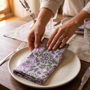 Puede incluir: Una servilleta de tela doblada con un patrón floral repetitivo en tonos morado, verde y blanco. La servilleta está colocada sobre un plato de cerámica de color blanco roto con motas, parte de un servicio de mesa con tenedor y vaso.