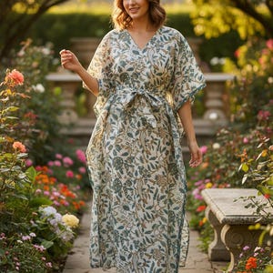 May include: A cream-coloured, long, flowing caftan with a V-neck and a tie-waist. The caftan features a floral pattern in teal and brown. The model is wearing the caftan in a garden setting with a stone bench and colourful flowers.