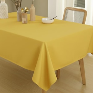 Puede incluir: Un mantel rectangular amarillo brillante cubre una mesa de madera. Un cuenco blanco con una cuchara negra, jarrones y utensilios están sobre la mesa. El mantel es de tela lisa y tiene un ligero brillo.