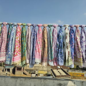 May include: A collection of colorful, patterned scarves hanging on a metal rod against a blue sky. The scarves display various floral and geometric designs in shades of blue, pink, green, and white, creating a vibrant display.