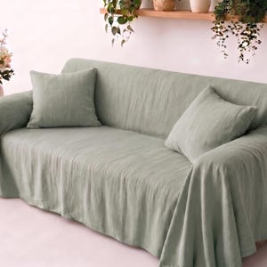 Könnte beinhalten: Ein salbeigrüner Sofabezug, der über eine Couch drapiert ist und eine strukturierte Stoffoberfläche aufweist. Das Set enthält zwei passende quadratische Wurfkissen in derselben salbeigrünen Farbe für ein einheitliches Erscheinungsbild.
