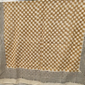 May include: A tan and white checkered textile with a black and white striped border. The fabric is hanging, showcasing a repeating square pattern in a grid-like arrangement. The border features vertical stripes and small geometric shapes.