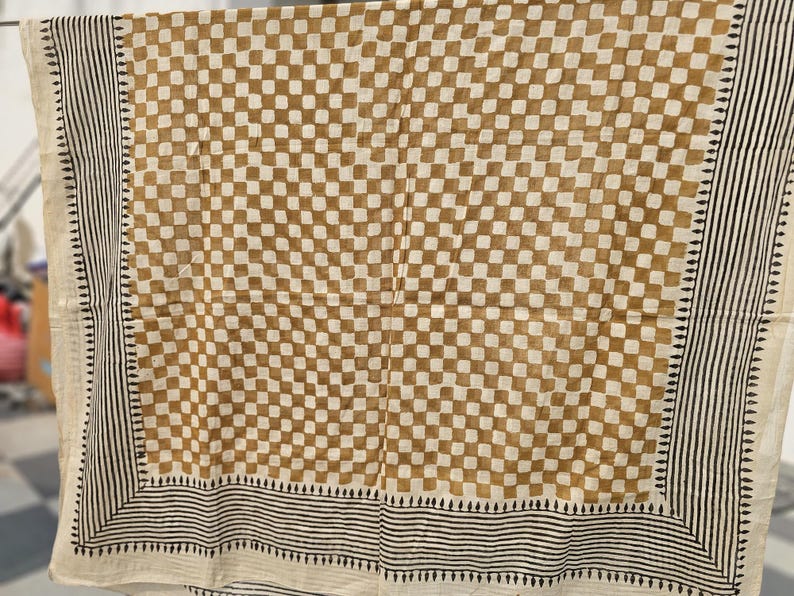 May include: A tan and white checkered textile with a black and white striped border. The fabric is hanging, showcasing a repeating square pattern in a grid-like arrangement. The border features vertical stripes and small geometric shapes.