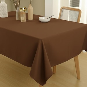 Op de afbeelding: Een effen bruin rechthoekig tafelkleed bedekt een tafel, met een hoek die naar beneden hangt. De tafel is gedekt met een witte kom, een lepel en decoratieve vazen. Het tafelkleed is gemaakt van een gladde, kreukvrije stof.