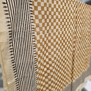 May include: A beige and brown patterned textile, possibly a scarf or shawl, hangs in the sunlight. The design features a checkered pattern in brown and beige, with vertical black stripes and leaf-like borders along the edges. The fabric appears lightweight.