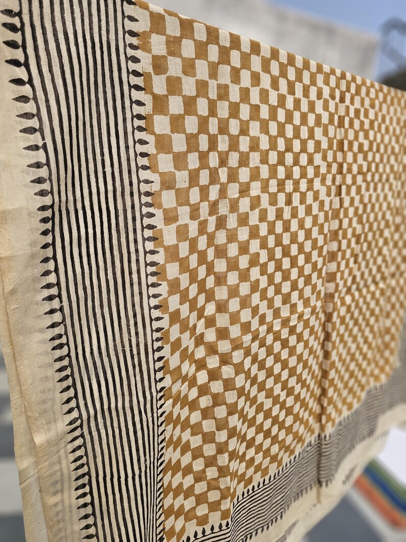 May include: A beige and brown patterned textile, possibly a scarf or shawl, hangs in the sunlight. The design features a checkered pattern in brown and beige, with vertical black stripes and leaf-like borders along the edges. The fabric appears lightweight.