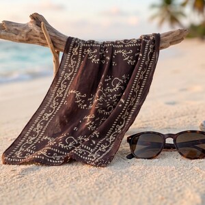 K&ouml;nnte beinhalten: Ein dunkelbraunes Bandana mit wei&szlig;em floralem Design h&auml;ngt an einem St&uuml;ck Treibholz an einem Sandstrand. Eine Schildpatt-Sonnenbrille liegt neben dem Bandana auf dem Sand. Der Hintergrund zeigt das Meer und Palmen.