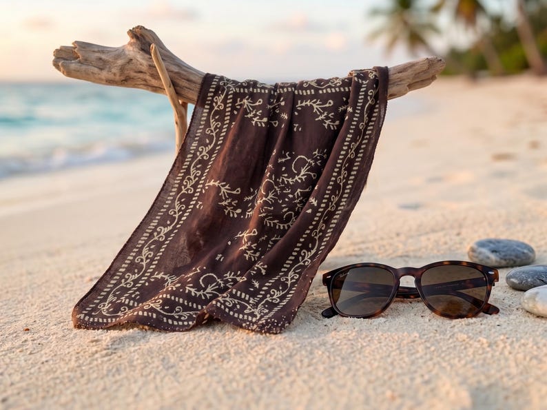 K&ouml;nnte beinhalten: Ein dunkelbraunes Bandana mit wei&szlig;em floralem Design h&auml;ngt an einem St&uuml;ck Treibholz an einem Sandstrand. Eine Schildpatt-Sonnenbrille liegt neben dem Bandana auf dem Sand. Der Hintergrund zeigt das Meer und Palmen.