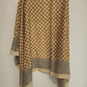 May include: A tan and cream-colored scarf with a checkered pattern. The scarf has a border with black and cream-colored stripes and a decorative edge. The scarf is hanging against a white wall.