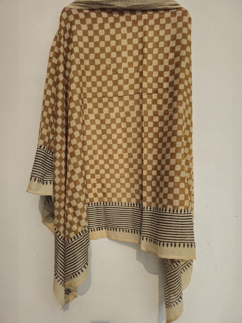 May include: A tan and cream-colored scarf with a checkered pattern. The scarf has a border with black and cream-colored stripes and a decorative edge. The scarf is hanging against a white wall.