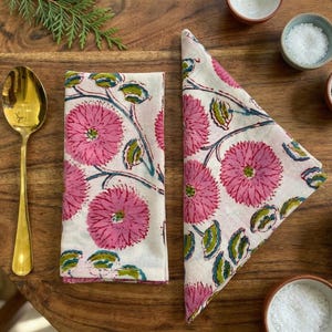 May include: Two white napkins with a pink floral design, a gold spoon, and small bowls of salt and pepper. The napkins display large pink flowers with green leaves and stems. The items are arranged on a wooden table.