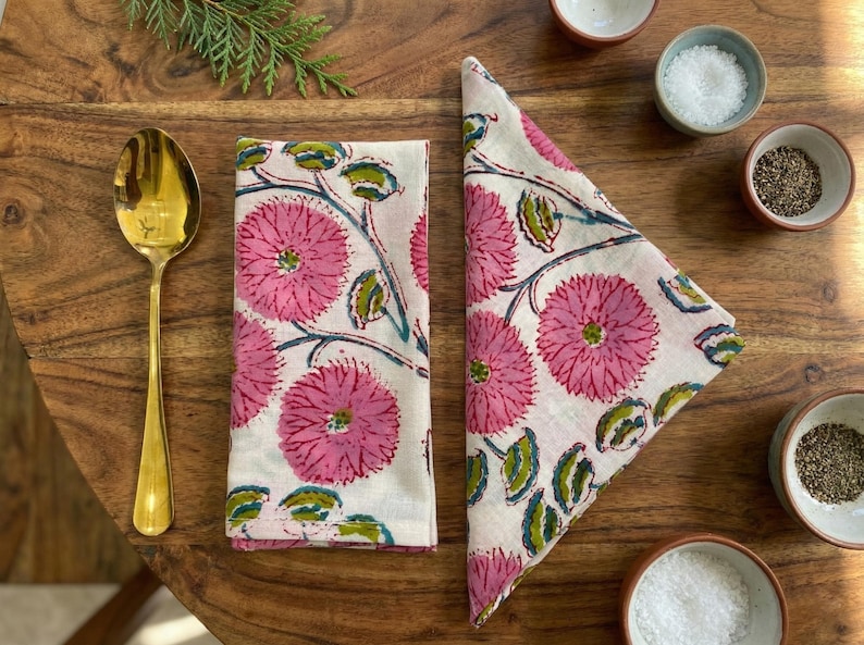 May include: Two white napkins with a pink floral design, a gold spoon, and small bowls of salt and pepper. The napkins display large pink flowers with green leaves and stems. The items are arranged on a wooden table.